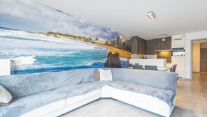 Een moderne woonkamer met een grote zeemuur, een grijze hoekbank en een minimalistische keuken met witte kasten en een inbouwoven.