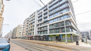 Een modern meerlaags gebouw met een commerciële ruimte op de begane grond, een tramlijn en een tramhalte.