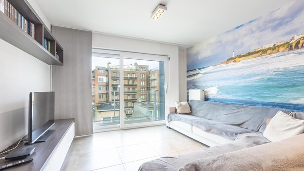 Een moderne woonkamer met een groot raam, een grijze bank, een flatscreen televisie en een schilderij van een kustlandschap.