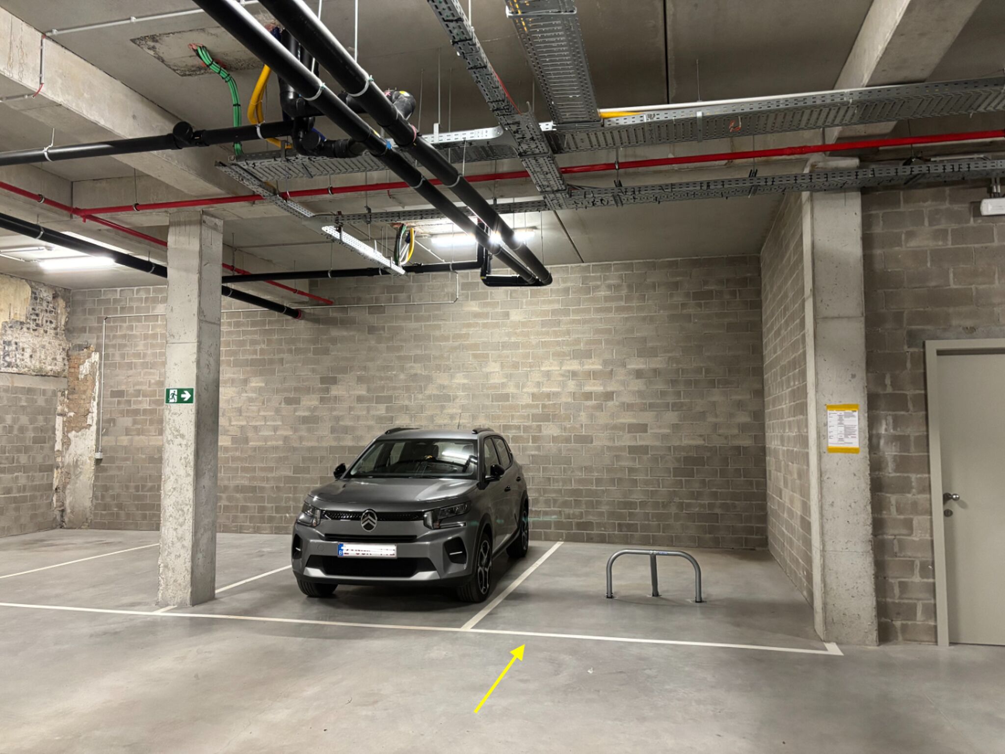 A parking garage with a car, concrete pillars, brick walls, and overhead pipes.