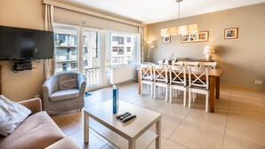 A modern living room with a sofa, a coffee table, and a TV. There is a dining area with a table and chairs. The room has large windows and a view of buildings outside. - Cosy flat near the centre (Koksijde)