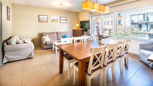 A modern living room with a wooden dining table, white chairs, a sofa, and a TV. - Cosy flat near the centre (Koksijde)