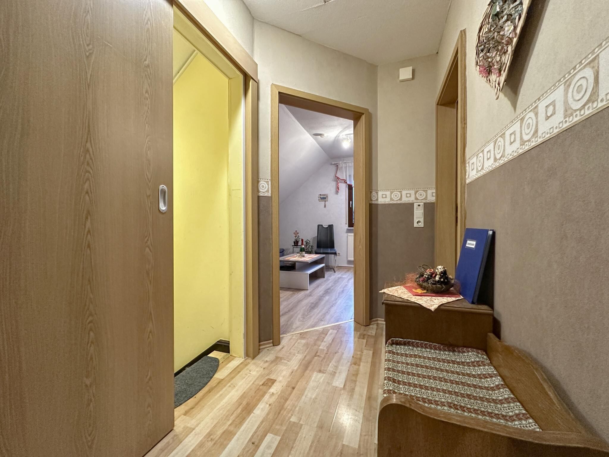A hallway with wooden flooring, a yellow door, and a small seating area with a patterned rug.