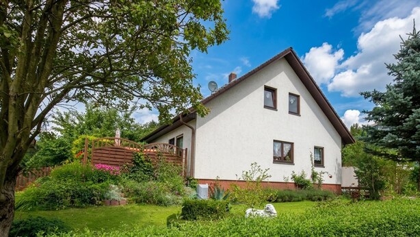 Ein zweistöckiges Haus mit weißer Fassade, einem geneigten Dach und einem kleinen Garten mit Hecke und Statue.