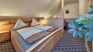 Ein Schlafzimmer mit schrägen Wänden, einem Bett mit zwei Kissen, einem Nachttisch mit Lampe und einer Pflanze.