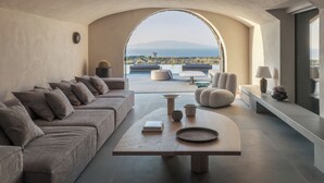 A modern living room with a sofa, armchairs, a coffee table, and a view of the outdoors through an arched window. - Charisma Estate Villa (Oia)