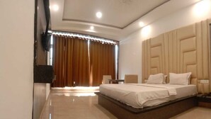 A modern hotel room with a large bed, a flat-screen TV mounted on the wall, and a view of the outdoors through a window with curtains.