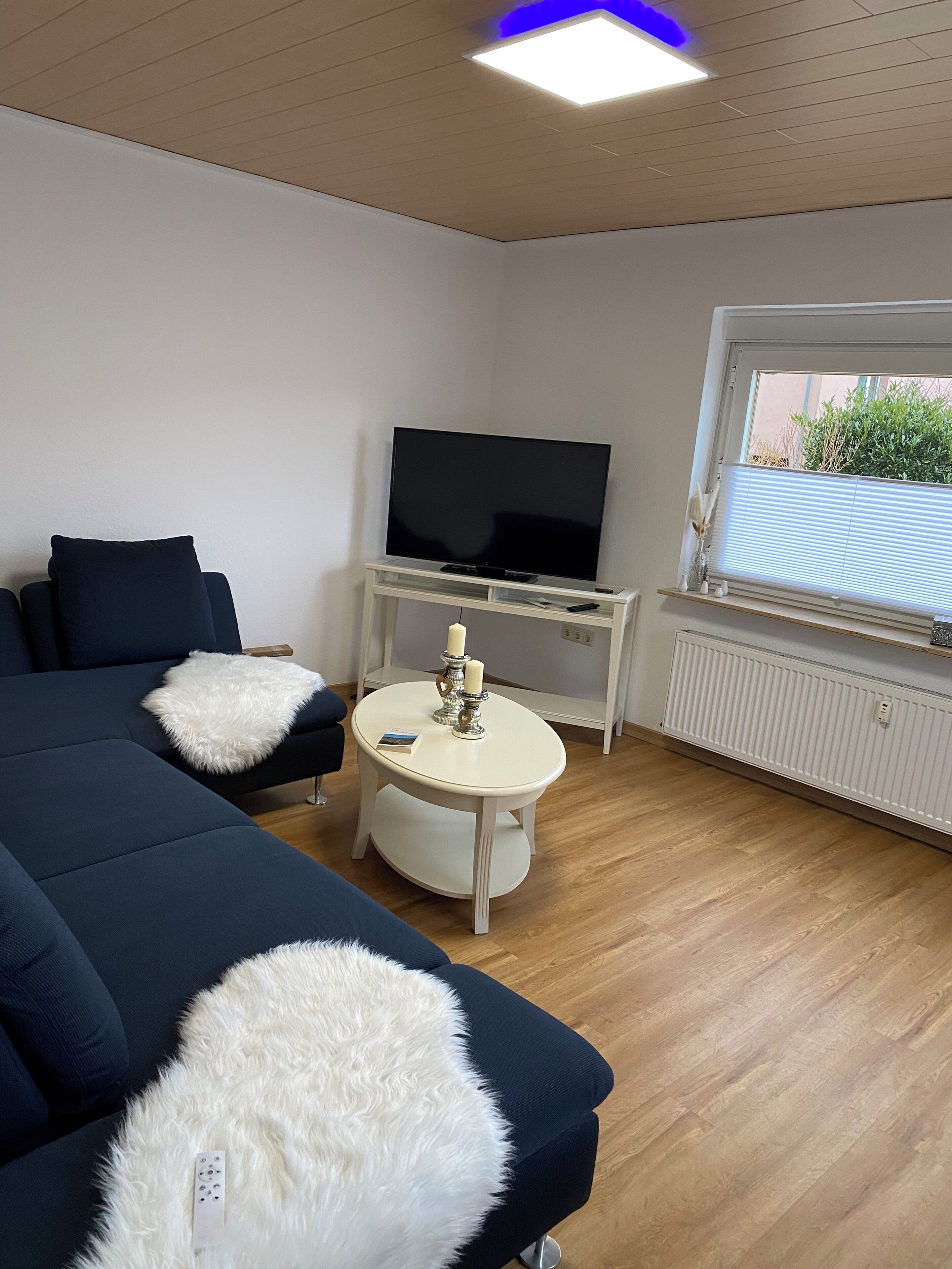 Un salon moderne avec un grand canapé de couleur foncée, une table ronde blanche ornée de bougies, un téléviseur à écran plat posé sur un meuble blanc et une fenêtre avec des stores.