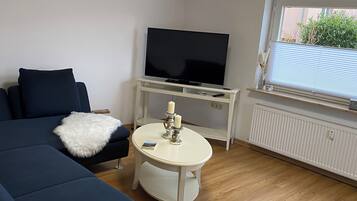Un salon moderne avec un grand canapé de couleur foncée, une table ronde blanche ornée de bougies, un téléviseur à écran plat posé sur un meuble blanc et une fenêtre avec des stores.