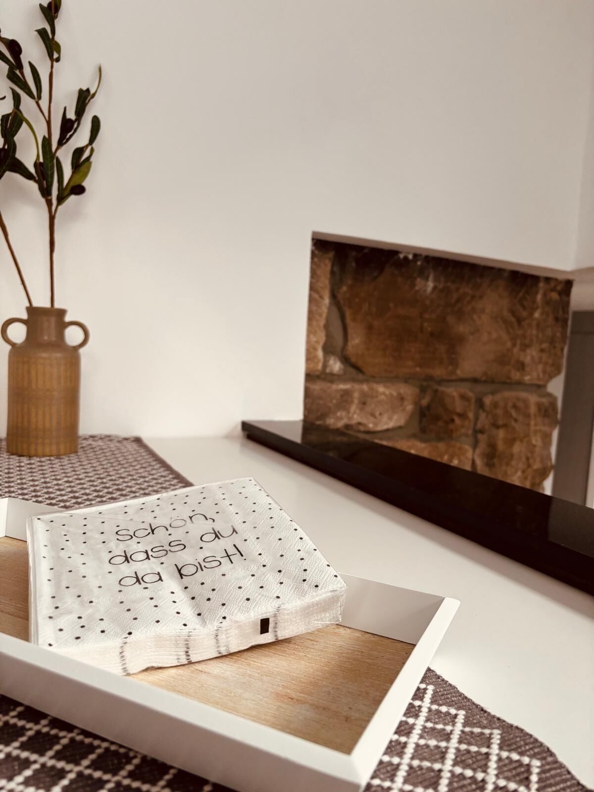Une œuvre d’art encadrée représentant un mur de pierres, un vase blanc avec une plante et un plateau blanc recouvert d’une nappe à pois.