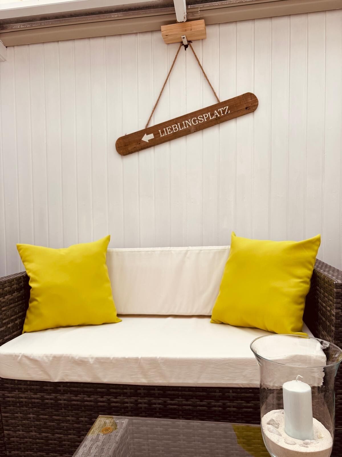 A wicker sofa with yellow cushions, a glass candle holder with a white candle, and a wooden sign above the sofa.