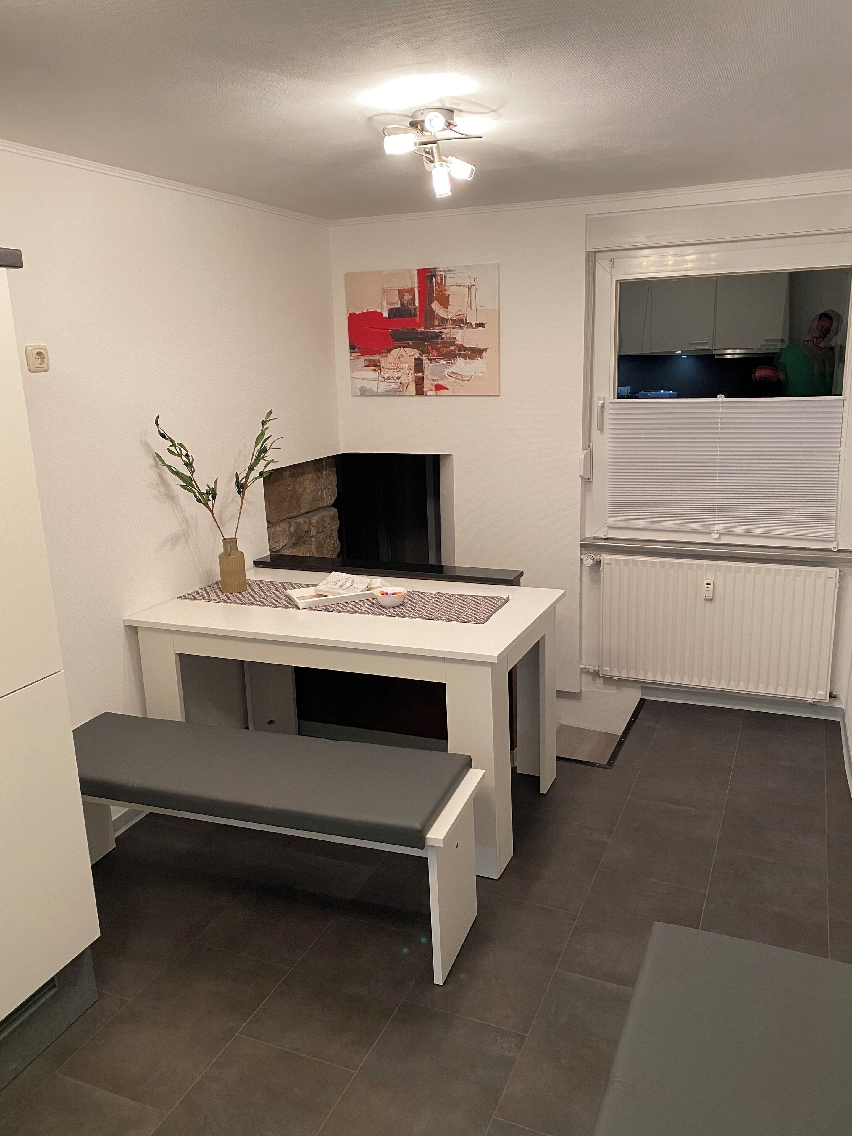 Een kleine keuken met een wit aanrecht, een bankje, een vaas met groen en een plafondlamp.