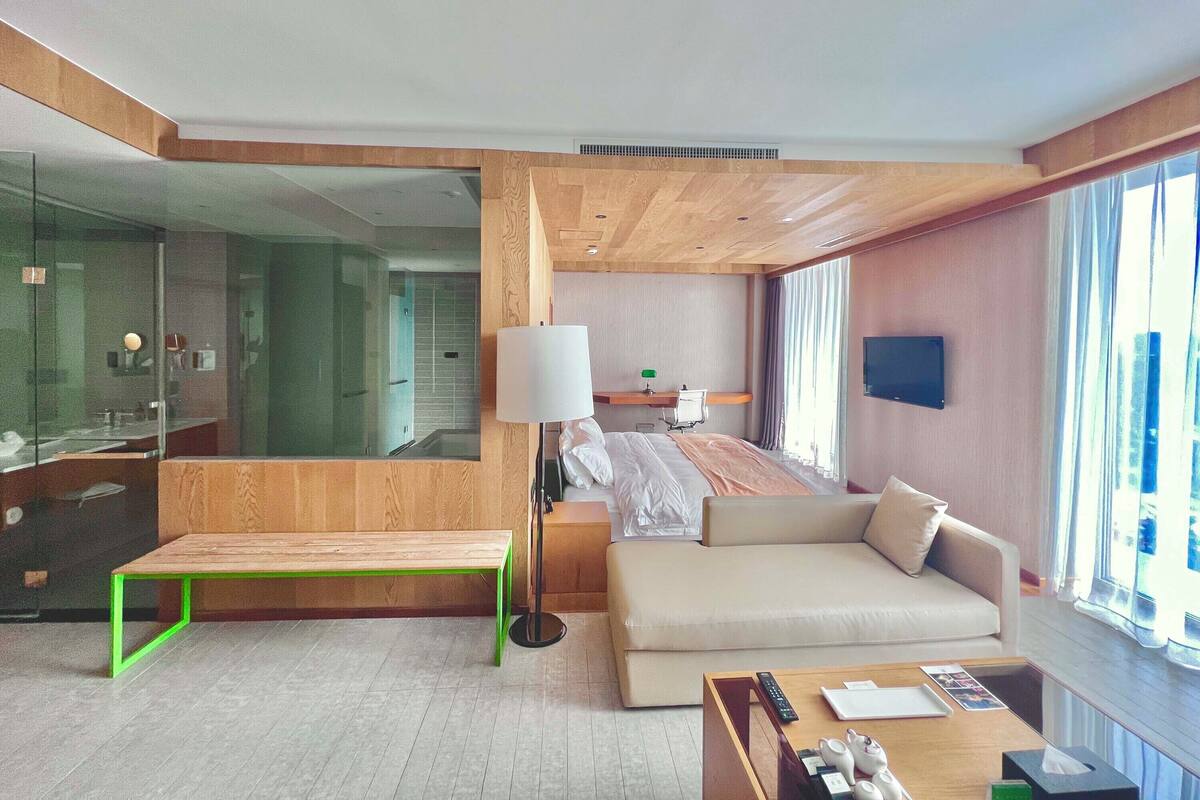 A modern hotel room with a large bed, a sofa, a bench, a bedside table, a lamp, and a TV.