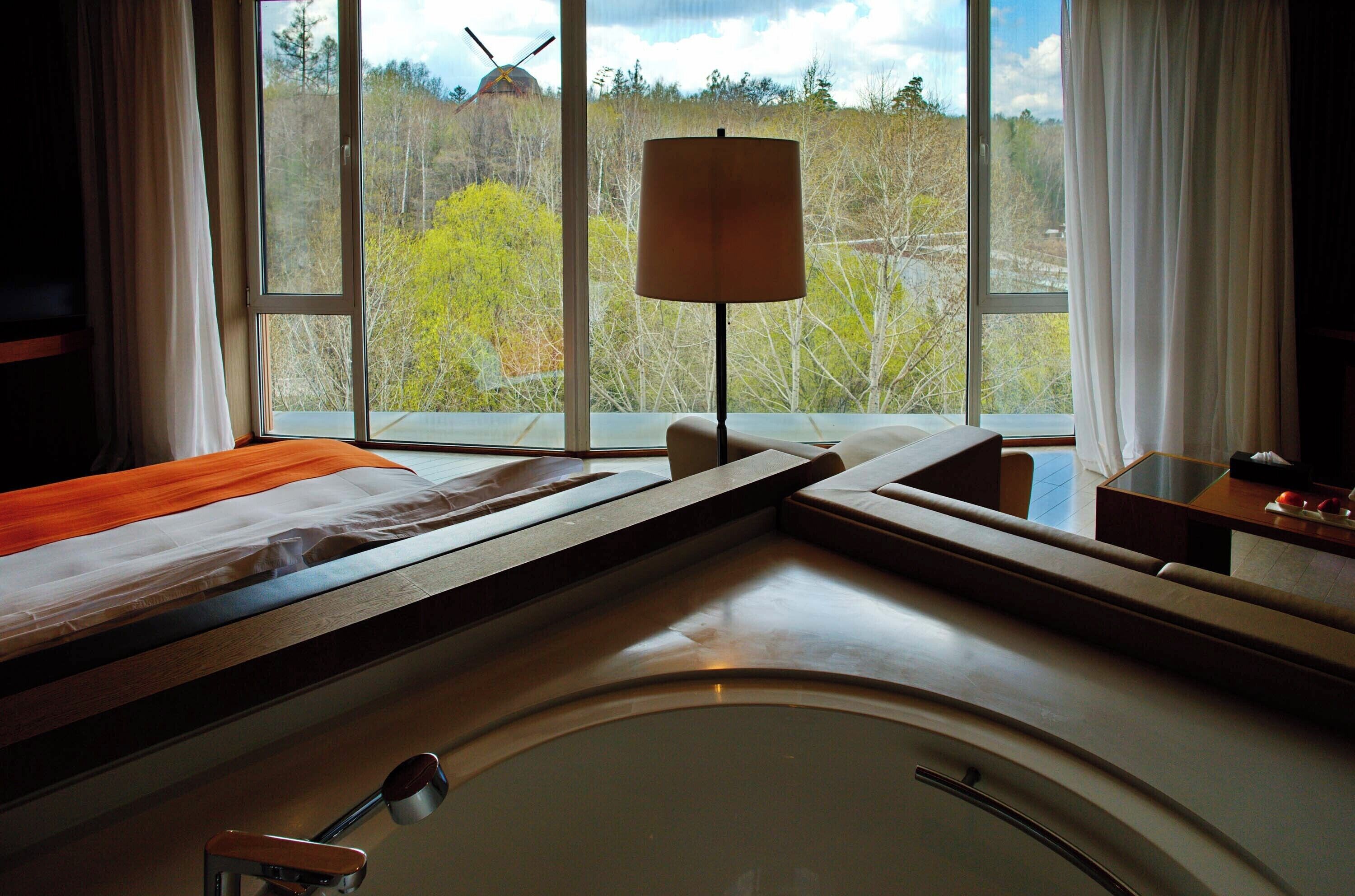 A hotel room with a bed, a bedside table, a lamp, and a view of a windmill and trees.