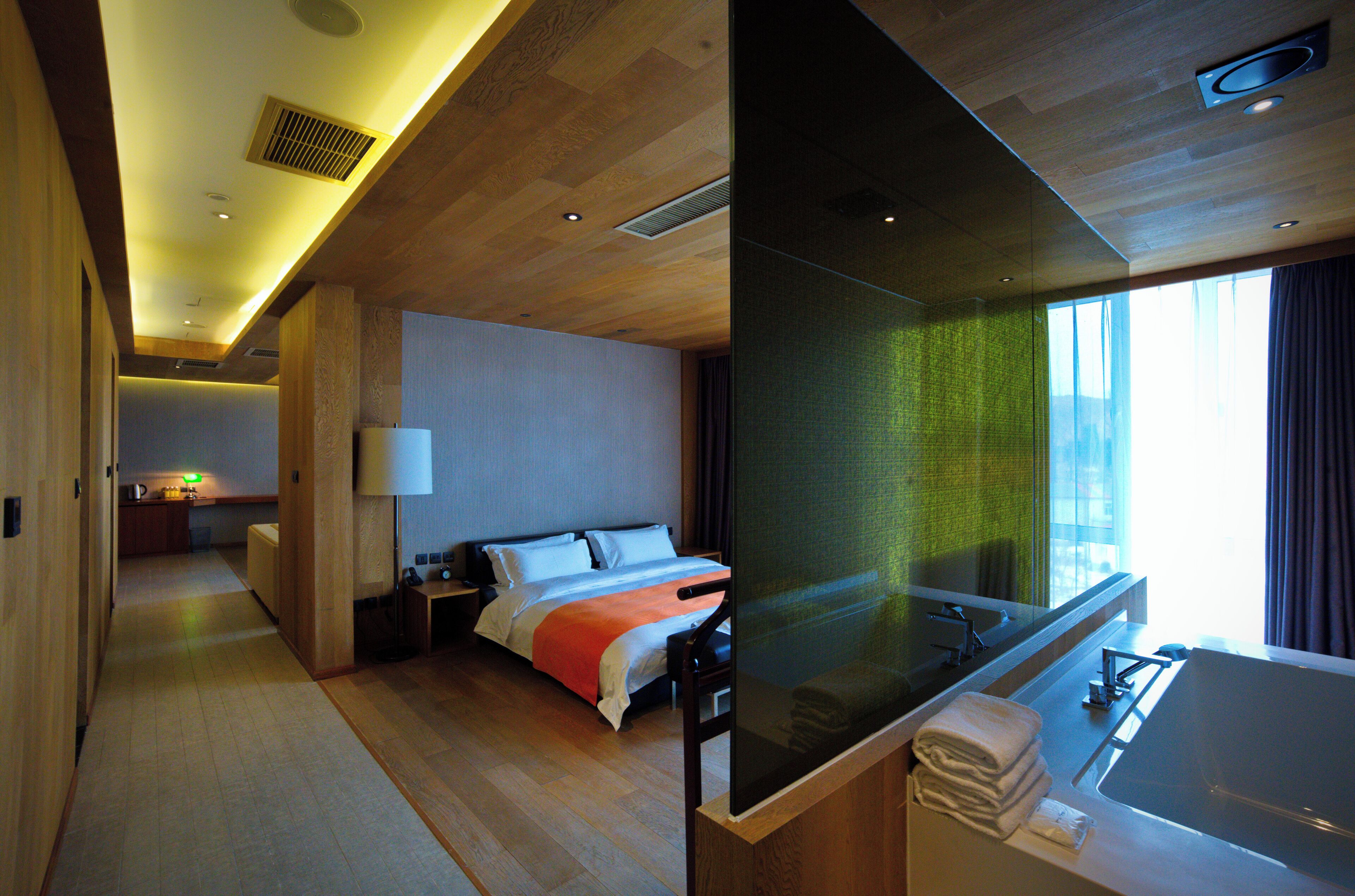 A modern hotel room with a large bed, a separate bathroom with a bathtub, and a view of the city through large windows.