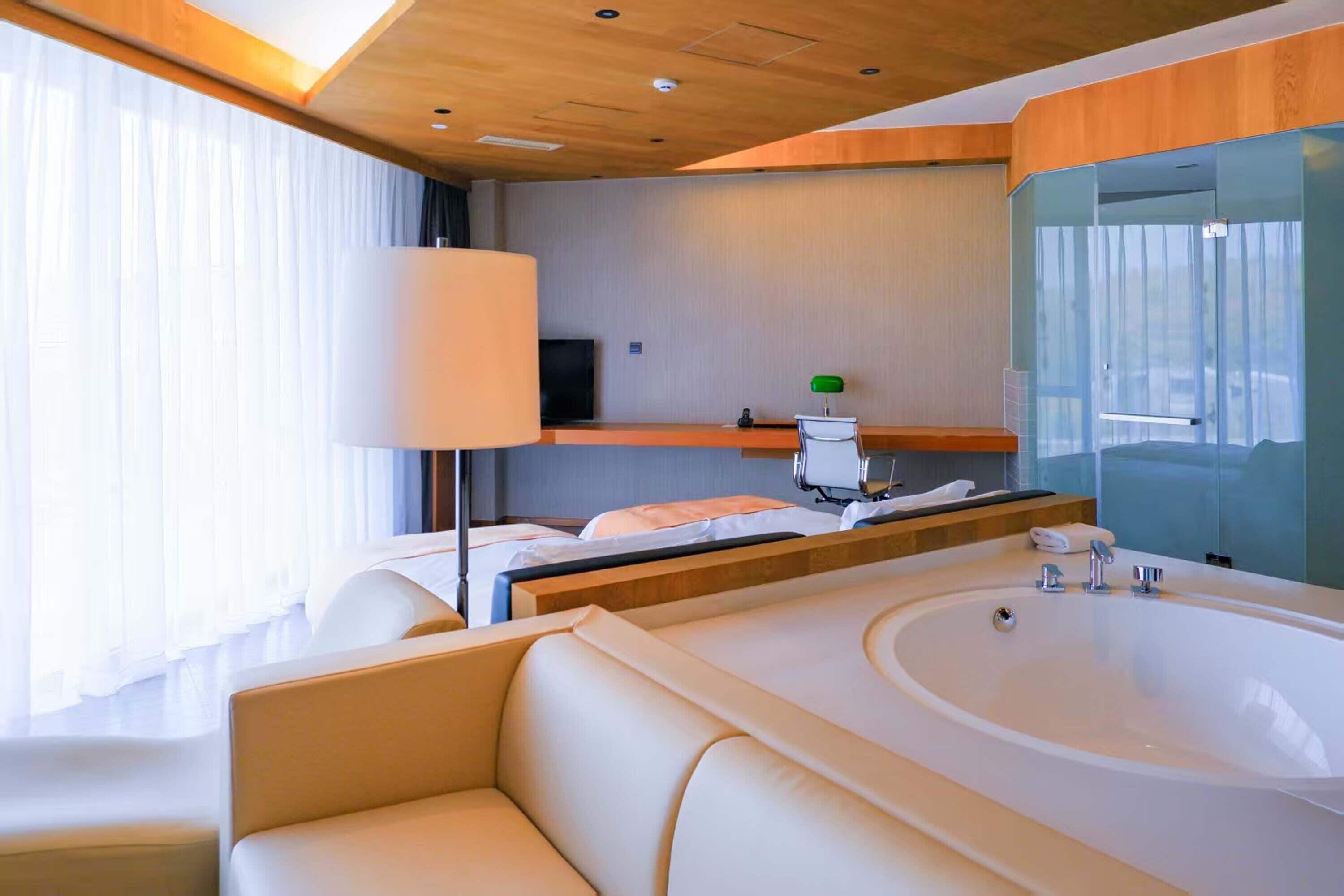 A modern hotel room with a large bathtub, a sofa, a flat-screen TV, and a large window with curtains.