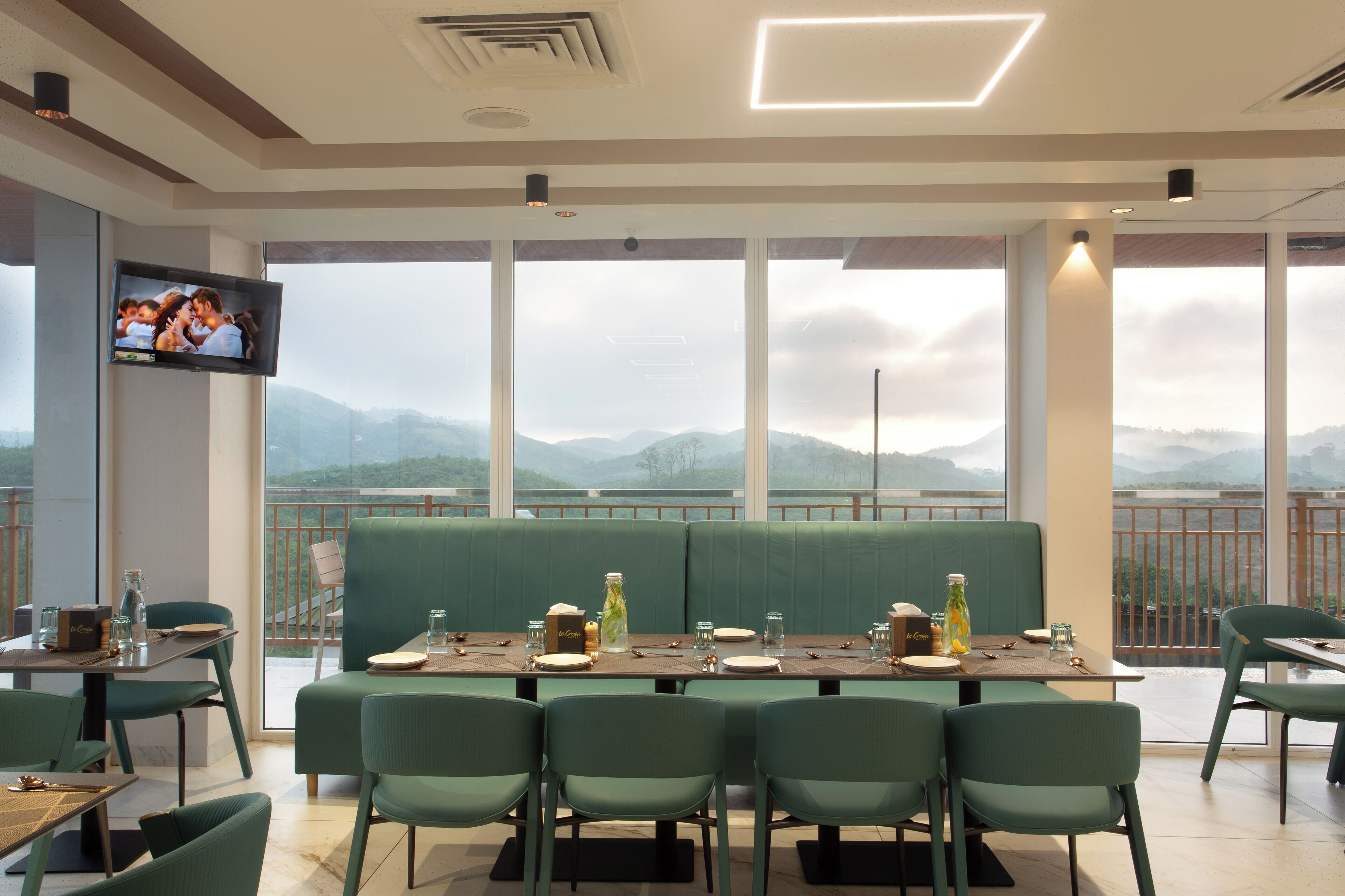 A restaurant with a large window view of mountains, green seating, and a flat-screen TV on the wall.