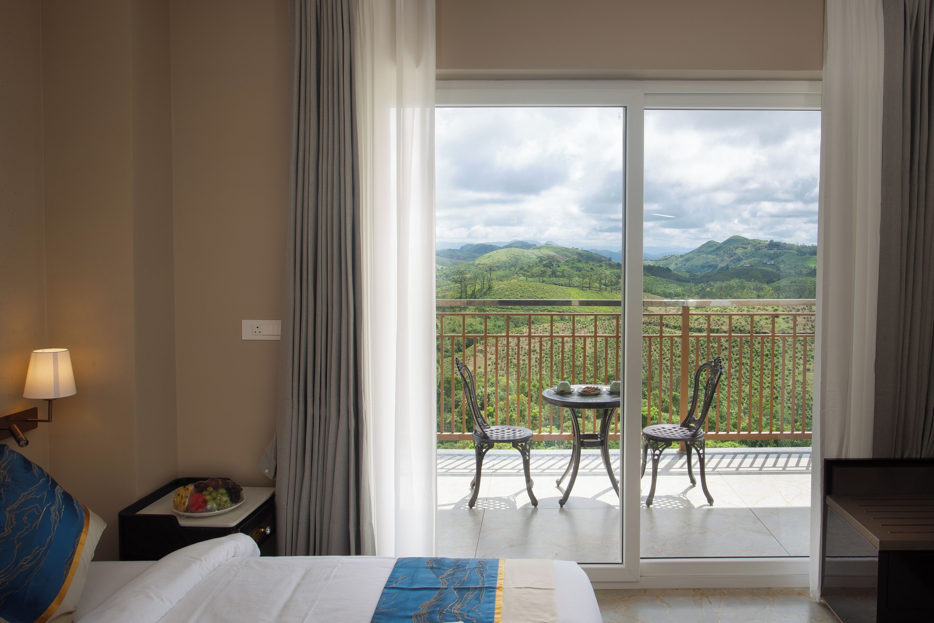Una habitación de hotel con cama, mesita de noche con lámpara, mesa de comedor con sillas y balcón con vistas a colinas verdes.