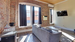 A living room with a brick wall, a sofa, a TV, and a window with curtains. - Walk to Courthouse Sq! Loft-Style Marshall Studio (Marshall)