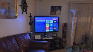 Ein Wohnzimmer mit einer Ledersofa, einem Glastisch mit Dekorationselementen, einem Flachbildfernseher, der eine Streaming-Oberfläche zeigt, einer Wanddekoration und einem gerahmten Bild über dem Fernseher.