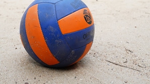 Een volleybal op een zandig oppervlak, met een gebouw en palmbomen op de achtergrond.