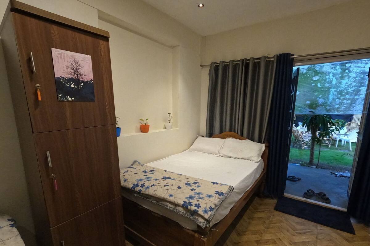 A bedroom with a wooden wardrobe, a bed with a floral bedspread, a small shelf with plants, and a view of the outdoors through a window with curtains.