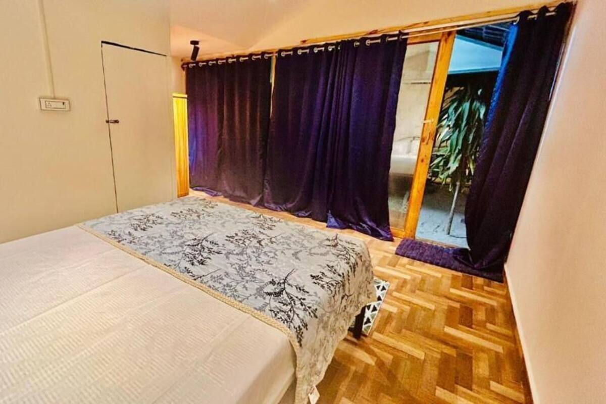 A bedroom with a bed, a window with curtains, a wooden floor, and a door.