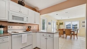Una cocina con armarios blancos, microondas, estufa y una zona de comedor con sillas y una mesa de madera.