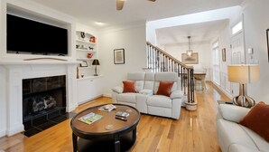 A spacious living room with a fireplace, a large flat-screen TV mounted above it, a white sofa with orange cushions, a wooden coffee table, and a staircase leading to an upper level. - Augusta Getaway Near Masters Porch Swing Deck (Augusta)
