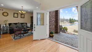 A spacious living room with a wooden floor, a sofa set, a chandelier, and a door leading to a covered porch with a view of a river and trees. - Luxury Ann Arbor Stay Theater Gym Game Room Spa (Ann Arbor)