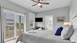 A bedroom with a large bed, a ceiling fan, a TV, and a balcony with a view of houses and trees. - Walk to Beach Pool Ocean View Parking Bunk Beds (Holly Ridge)