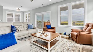 A living room with a beige sofa, a brown leather armchair, a wooden coffee table, and a large window with a view of a fence. - Walk to Beach Pool Ocean View Parking Bunk Beds (Holly Ridge)