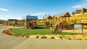 Ein Spielplatz mit verschiedenen Spielgeräten, einer überdachten Sitzgelegenheit und einem Gebäude mit Satteldach.