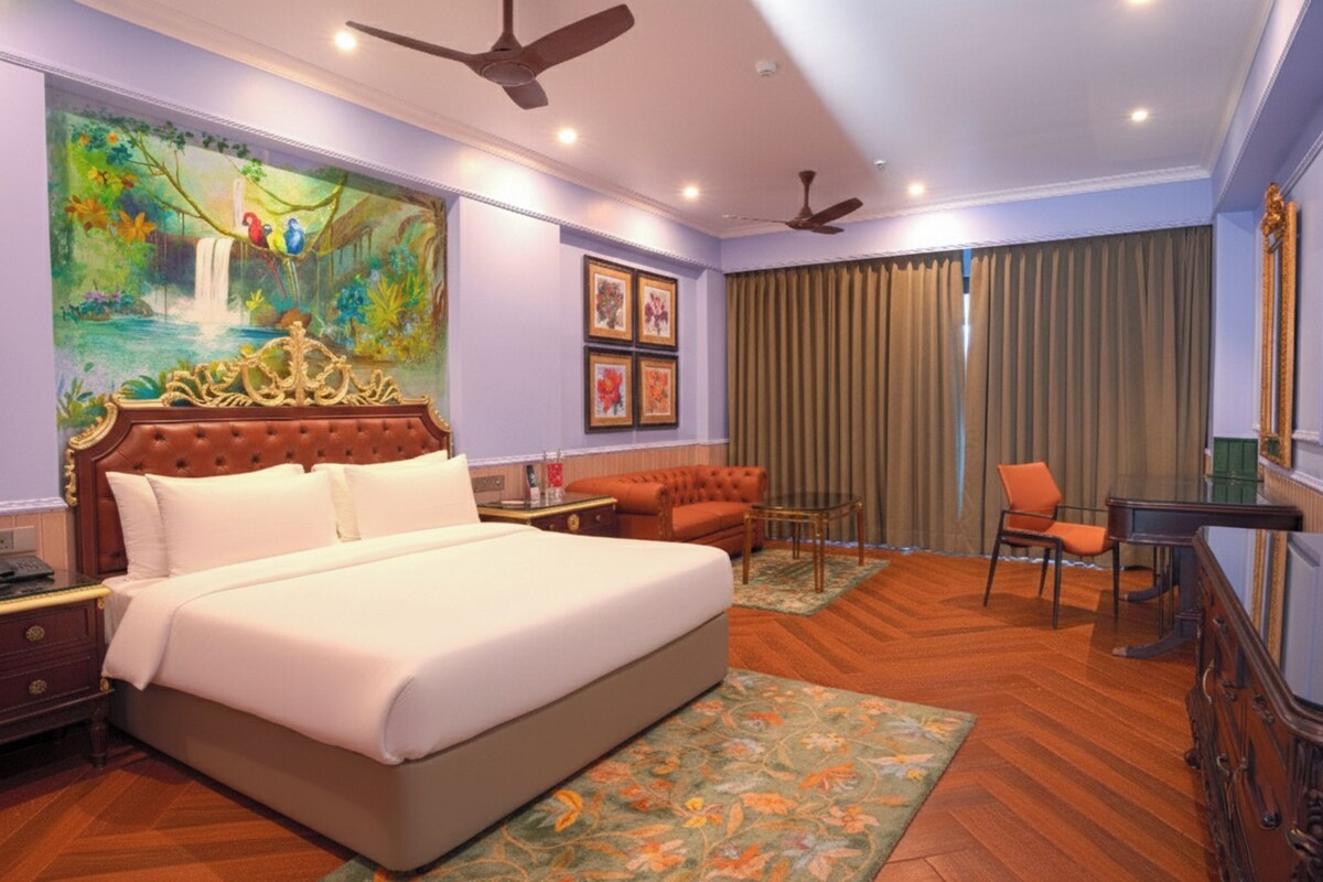 A hotel room with a large bed, a desk, a chair, a ceiling fan, and a painting on the wall.