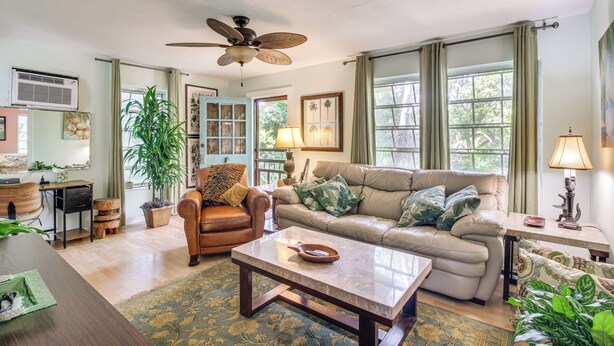 A spacious living room with a large sofa, a coffee table, and a ceiling fan. There is a large window with curtains, a potted plant, and a lamp on a side table. - Gateway to the Everglades! Tropical Jungle Cottage (Miami)