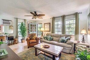 A spacious living room with a large sofa, a coffee table, and a ceiling fan. There is a large window with curtains, a potted plant, and a lamp on a side table. - Gateway to the Everglades! Tropical Jungle Cottage (Miami)