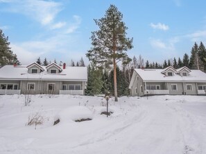Een woongebied met twee huizen, elk met een puntige dakschild en een veranda, omgeven door besneeuwde grond en bomen.
