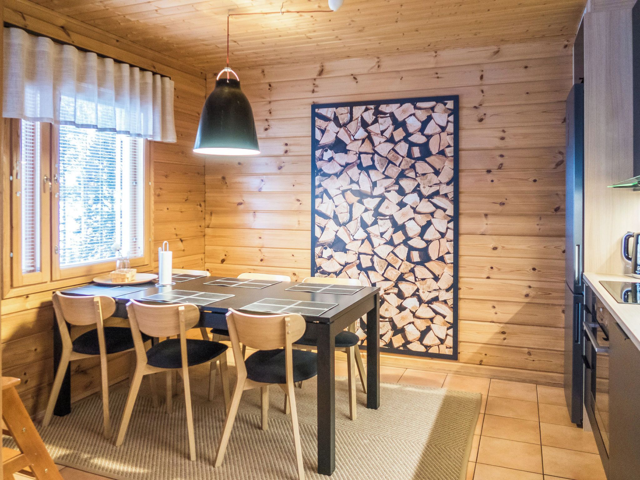 Ein Holzinnenraum mit einem Esstisch, Stühlen, einer hängenden Leuchte und einer Wanddekoration aus Holzscheiten.