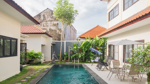 A swimming pool surrounded by a garden with chairs and a table, a statue, and a building with a red-tiled roof. - Samurai Sanur Hotel (Denpasar)