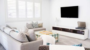 A living room with a sofa, ottoman, and a flat-screen TV mounted on the wall. - Waterfront - Kolora Lake House - Oceanstays (Yamba)