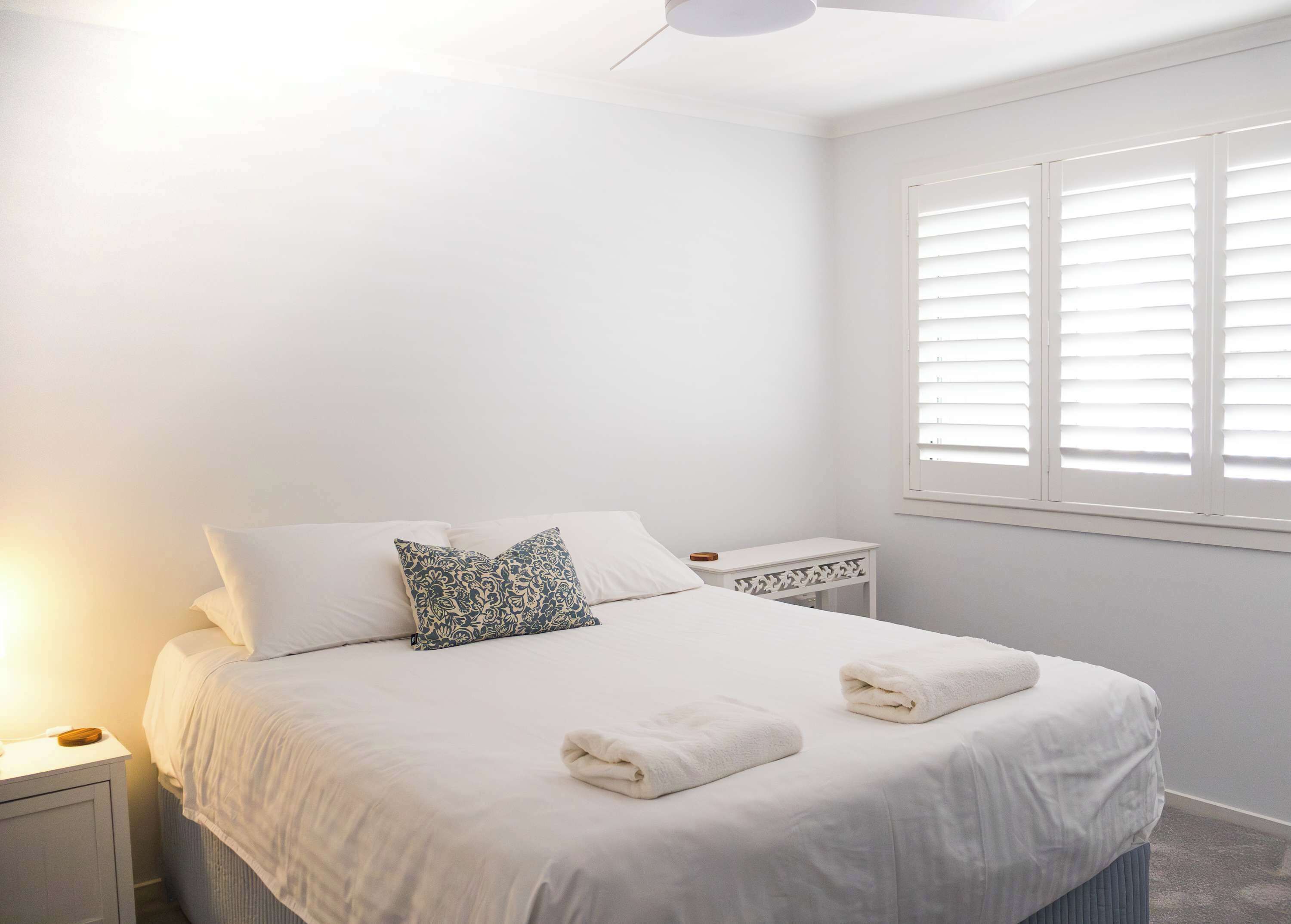 A neatly made bed with white linens and two folded towels, a bedside table with a lamp, and a window with blinds.