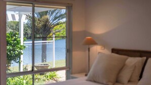 A bedroom with a bed, a nightstand, a lamp, and a view of a body of water through a sliding glass door. - Waterfront - Kolora Lake House - Oceanstays (Yamba)