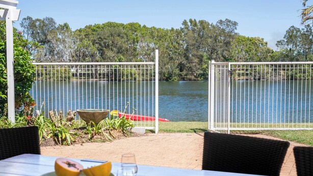A table with a plate of fruit and a glass of water, a white fence, and a view of a lake with trees in the background. - Waterfront - Kolora Lake House - Oceanstays (Yamba)