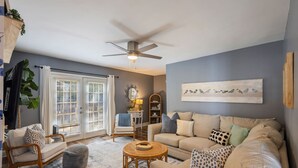 A living room with a beige sofa, a round coffee table, a wooden chair, and a large window with blinds. - Hot Tub BBQ Retreat Spacious Backyard Pet-friendly (North Augusta)