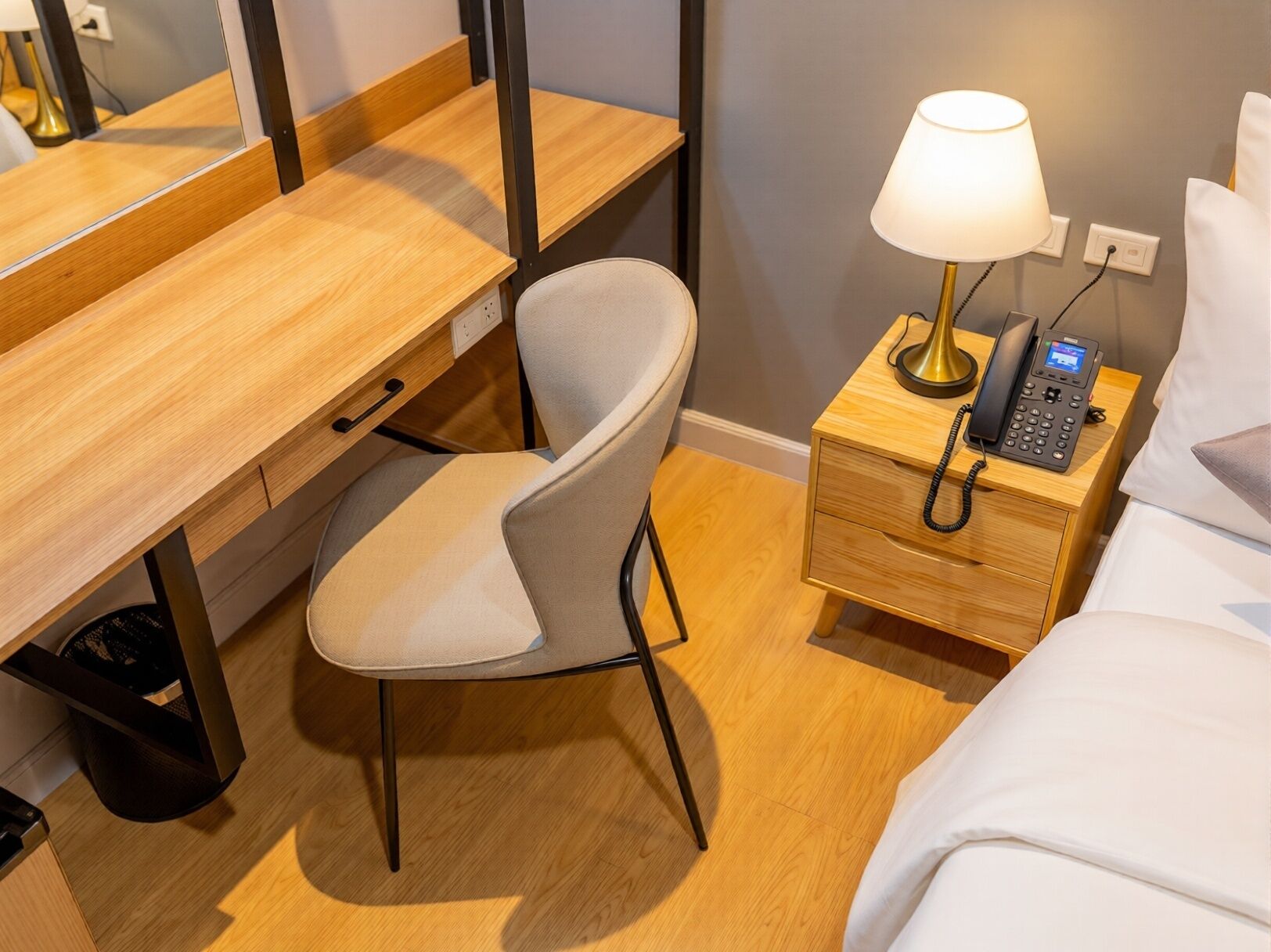 A hotel room with a wooden desk, a chair, a bedside table with a lamp, a phone, and a bed.