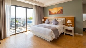 A modern hotel room with a large bed, a bedside table with a lamp, a view of the city through floor-to-ceiling windows, and a balcony with outdoor furniture. - Doña Manor Boutique Hotel (Olongapo)