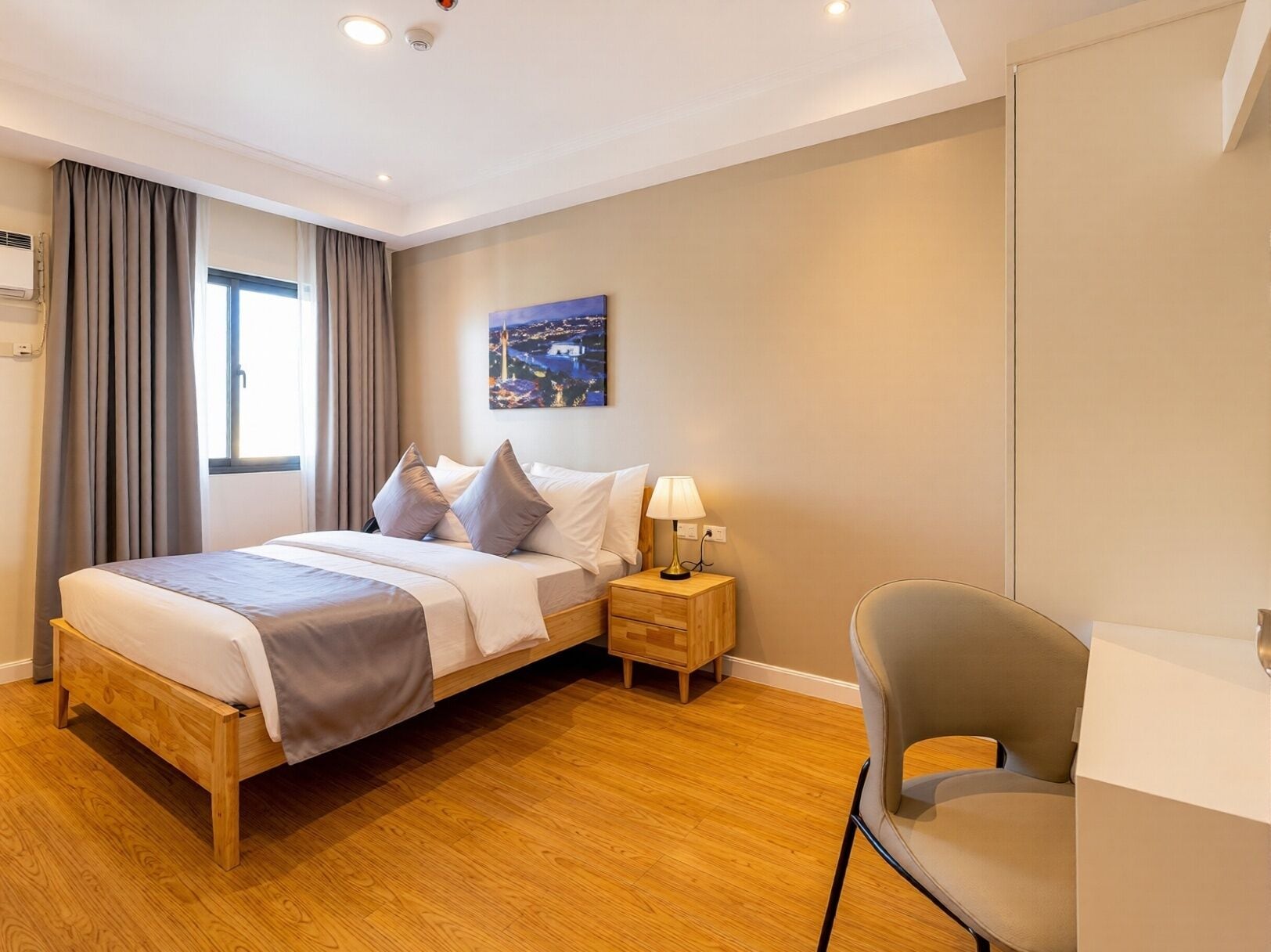 Une chambre d’hôtel avec un lit en bois, une table de chevet avec une lampe, une chaise grise et un tableau représentant un paysage urbain.
