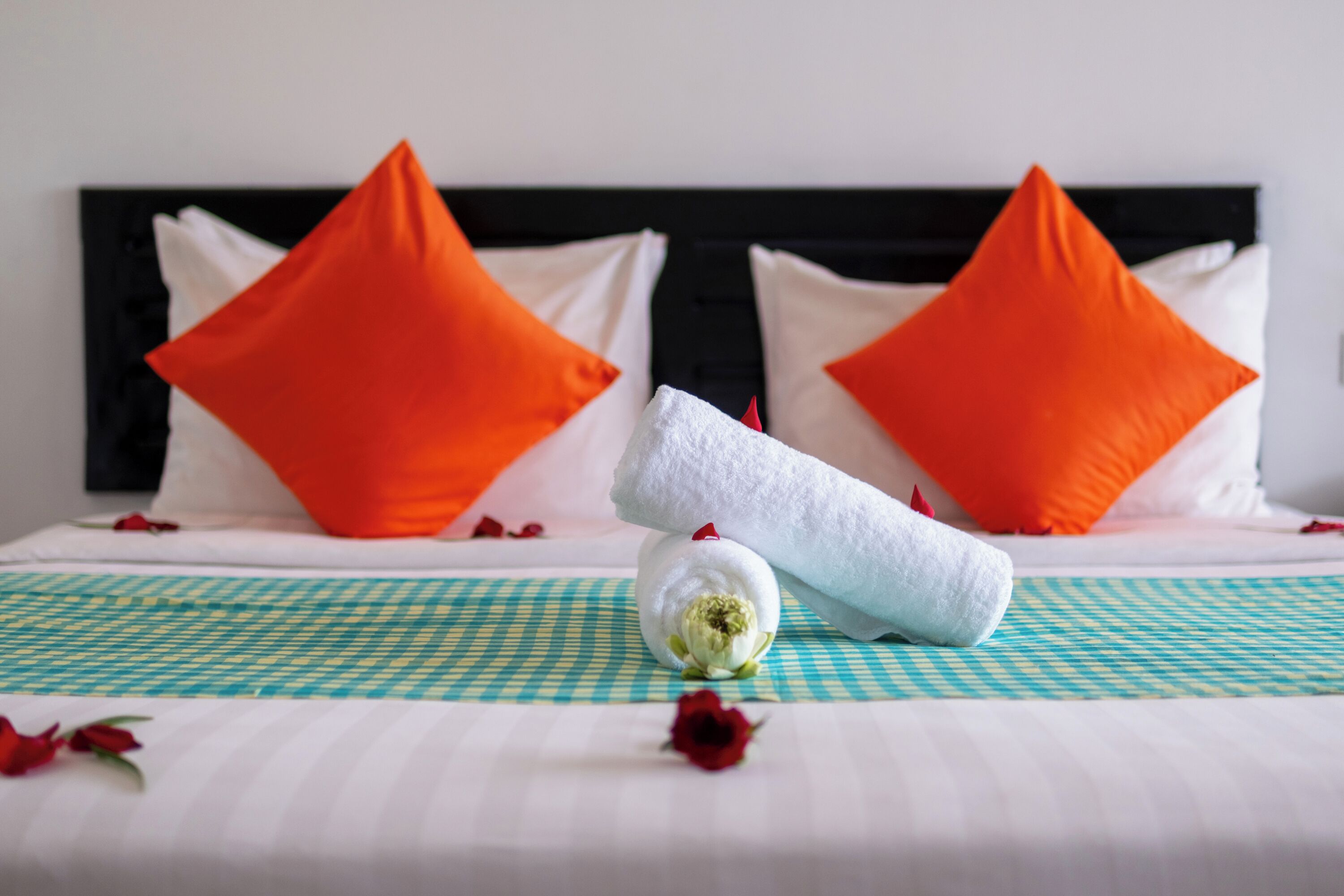 Un lit bien fait, agrémenté d’oreillers orange, d’une serviette blanche roulée et d’un arrangement floral décoratif au centre.