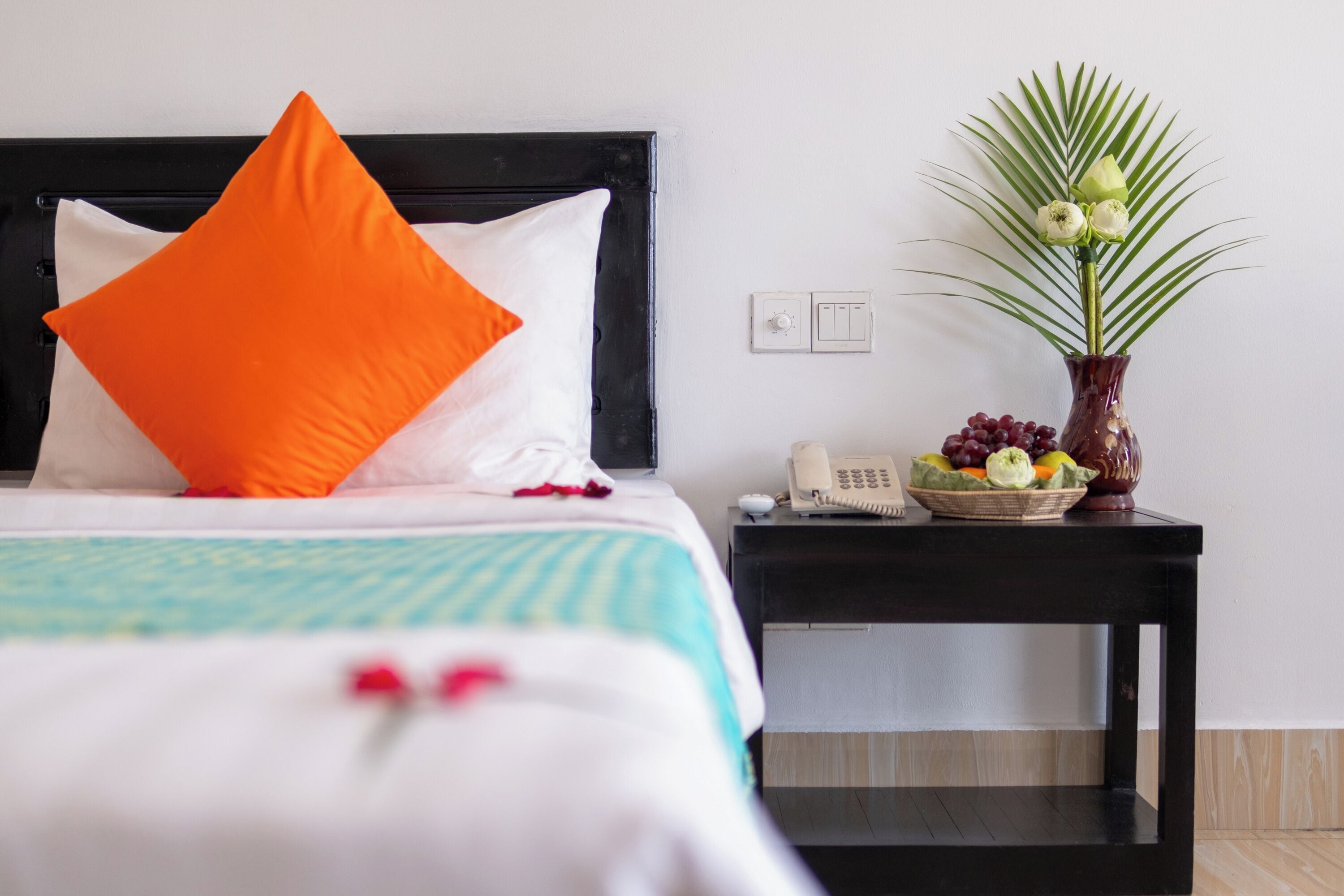 A neatly made bed with an orange pillow, a bedside table with a phone, a vase with flowers, and a fruit basket.