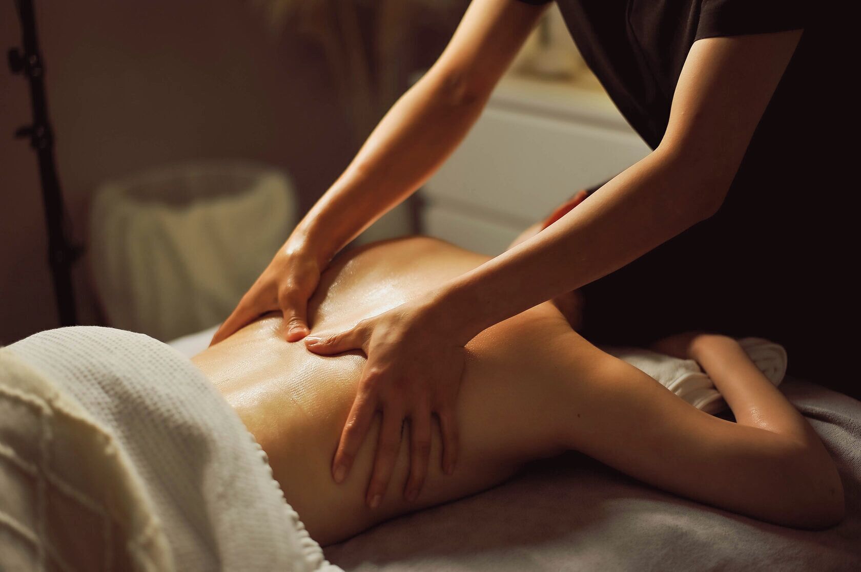 A person receiving a back massage on a bed in a room with a window and a lamp in the background.
